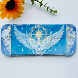 May include: A blue and white Switch console case featuring a celestial design with angel wings, stars, and a compass rose. The case has a glossy finish and is decorated with glitter accents. A sunflower and greenery are in the background.