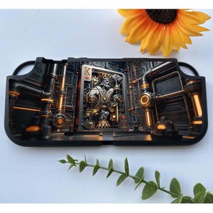 May include: Black and gold gaming console case featuring a steampunk-style design with a playing card image of the King of Hearts. The case has a detailed industrial background with pipes and gears. A yellow sunflower is in the background.