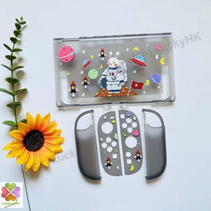 May include: A translucent gray protective console case and controller covers featuring a space-themed print with a rabbit astronaut, rockets, planets, and stars. The design includes a red flag and various celestial bodies scattered across the surface.