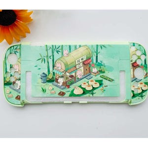 Puede incluir: Una funda verde claro para Nintendo Switch con una ilustración de dibujos animados de una tienda de conejos en un bosque de bambú. El diseño incluye alimentos y pequeños personajes de conejos. La funda tiene recortes para los botones y puertos de la consola.