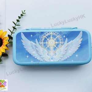 May include: A rectangular light blue pencil case featuring a graphic print of large white feathered wings centred around a glowing circular celestial emblem against a blue sky background with small white stars and a handle on the top edge.