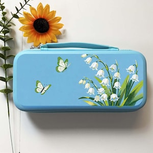 May include: A rectangular light blue carrying case featuring a printed design of white lily of the valley flowers with green leaves and two butterflies with green and white wings. The case has a matching light blue handle and a zipper closure.