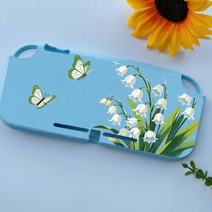May include: A light blue Switch case featuring a floral design of white lily of the valley flowers and green leaves. Two butterflies with green and white wings are also depicted. A yellow sunflower is in the background.