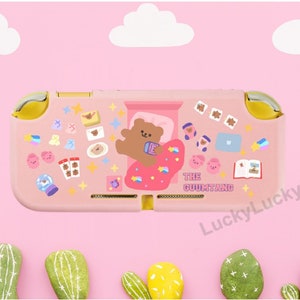 Può includere: Custodia rosa per Nintendo Switch con un orso marrone che indossa un pigiama e una coperta rosa e blu. La custodia è decorata con una varietà di adesivi, tra cui stelle, cuori e nuvole. Il testo "THE GUUMTANG" è stampato sulla custodia.