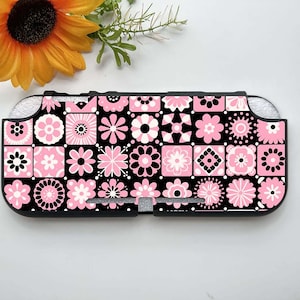 Puede incluir: Una funda negra y rosa con estampado floral para una consola de juegos portátil. La funda presenta una cuadrícula de flores rosas y blancas sobre un fondo negro. La funda está diseñada para ajustarse perfectamente al dispositivo.