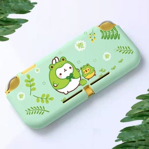 May include: A light green Nintendo Switch Lite case with a cartoon frog design. The case has a matte finish and features a large frog character in a frog hat, with two smaller frog characters. The design is complemented by floral and leaf accents.