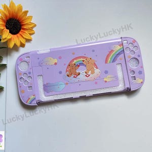 Pode incluir: Uma capa protetora para console na cor lavanda com um gráfico de dois ursos marrons sentados em uma nuvem sob um arco-íris. A capa inclui recortes para botões e joysticks, decorada com pequenas estrelas e nuvens. O texto LuckyLuckyHK é visível.