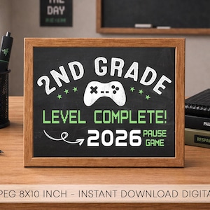 May include: A framed chalkboard sign with the text "2ND GRADE LEVEL COMPLETE! 2026 PAUSE GAME" in white and green. A video game controller graphic is centered above the text. The sign is on a wooden desk.