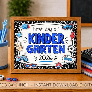 May include: A framed sign with a black and white speckled border featuring the text first day of KINDERGARTEN 2026 in blue. The design includes illustrations of a blue cap, race car, football, game controller, sunglasses, and fire.