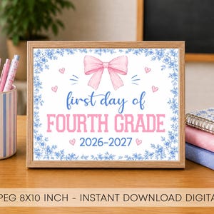 May include: A framed sign with a blue floral border, a pink bow, and the text first day of FOURTH GRADE 2026-2027. The sign is displayed next to a striped pen holder with various pens and a stack of notebooks on a wooden desk surface.