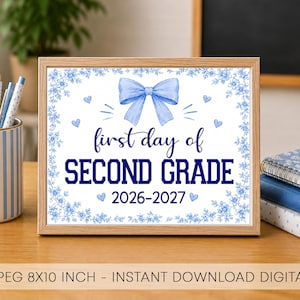 Può includere: Un cartello incorniciato con un fiocco azzurro, un bordo floreale e il testo "first day of SECOND GRADE 2026-2027". Il cartello è esposto su una scrivania accanto a un portapenne a righe blu e bianche e a una pila di quaderni blu.