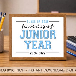May include: A rectangular wooden frame containing a white sign with black and light blue text that reads: CLASS OF 2028, first day of, JUNIOR YEAR, 2026-2027. The sign is displayed on a wooden desk next to a pencil holder and notebooks.