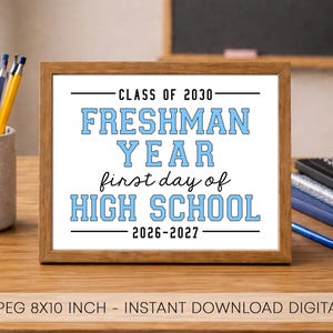 May include: A rectangular wooden frame containing a white sign with black and light blue text that reads: CLASS OF 2030 FRESHMAN YEAR first day of HIGH SCHOOL 2026-2027. The sign is 8x10 inches and is displayed on a wooden desk surface.