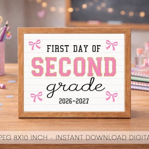 May include: A rectangular wooden frame containing a white sign with horizontal lines. The text reads FIRST DAY OF SECOND grade 2026-2027 in black and pink varsity-style lettering. Four small pink bows are positioned in the corners of the sign.