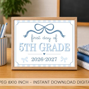 Könnte beinhalten: Ein gerahmtes Schild mit hellblau-weiß gestreiftem Hintergrund, einer blauen Schleifenillustration und dem Text: first day of 5TH GRADE 2026-2027. Das Schild steht auf einem Holztisch neben einem gestreiften Stiftehalter und gestapelten Notizbüchern.