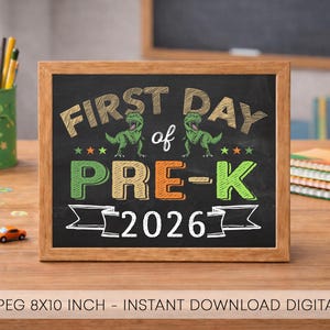以下が含まれることがあります： 明るい木製のフレームに入った長方形の黒板サイン。FIRST DAY of PRE-K 2026という文字がカラフルなチョーク風のフォントで描かれています。黒い背景に、2匹の緑色の恐竜のイラスト、小さな星、バナーのグラフィックがデザインされています。