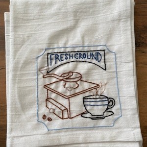 May include: White kitchen towel with blue and brown embroidery of a coffee grinder and a cup of coffee. The embroidery says "Fresh Ground".
