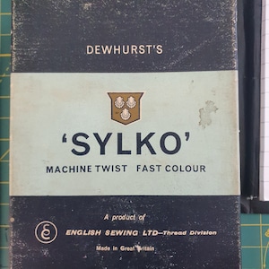 Puede incluir: Una caja negra y blanca con el texto "DEWHURST'S 'SYLKO' MACHINE TWIST FAST COLOUR" y un escudo con tres conchas. La caja también dice "A product of ENGLISH SEWING LTD-Thread Division Made in Great Britain."