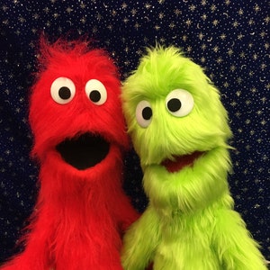 May include: Two fuzzy hand puppets, one red and one green, with large eyes and open mouths. They are both facing forward and appear to be smiling.