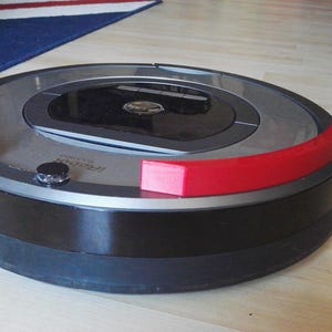 May include: A black and silver robotic vacuum cleaner with red bumper guards. The vacuum cleaner is on a wooden floor.