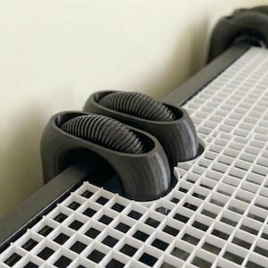 May include: Close-up of a black and white grid system with black rollers. The rollers are cylindrical with a ridged texture. The grid is white with a square pattern. The system is mounted on a wall.
