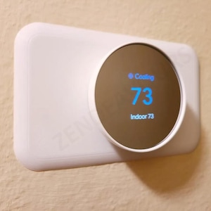 Puede incluir: Un termostato inteligente blanco montado en una pared beige. La pantalla digital muestra "Cooling" y la temperatura es de 23 grados Celsius. El termostato tiene una pantalla circular y una base rectangular.