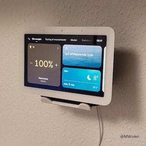 Peut inclure: Un appareil domotique blanc avec un écran tactile affichant l'heure, la température et une notification pour jouer des sons relaxants. L'appareil est monté sur un mur avec un cordon blanc.