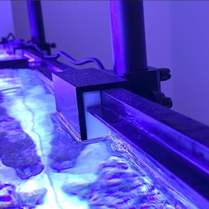 May include: A close-up of a black aquarium light fixture mounted to a black metal track. The light fixture has a white LED panel and is positioned over a blue-lit aquarium.