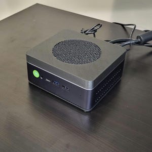 Puede incluir: Un ordenador negro de forma cuadrada con una rejilla de altavoz circular en la parte superior. El ordenador tiene un botón de encendido verde y varios puertos en el lateral.