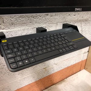 Op de afbeelding: Een zwart toetsenbord met een ingebouwd touchpad is met een zwarte beugel aan een muur gemonteerd. Het toetsenbord is met een zwarte kabel aan een computermonitor aangesloten.