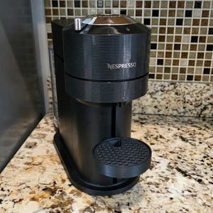 May include: A black Nespresso coffee machine with a round drip tray. The machine has a cylindrical design with horizontal ridges and the brand name "Nespresso" printed on the front. It sits on a speckled countertop.