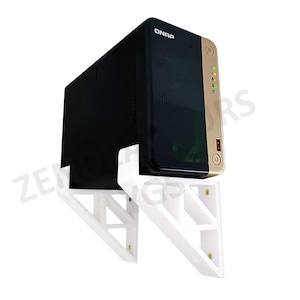 Puede incluir: Un dispositivo de almacenamiento conectado a la red (NAS) QNAP negro y dorado montado en un soporte blanco. El NAS tiene un panel lateral dorado con un puerto USB y un botón de encendido. El soporte blanco está montado sobre una superficie blanca.