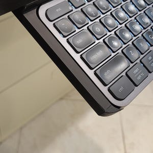 Puede incluir: Un teclado negro y plateado con teclas retroiluminadas. Las teclas están etiquetadas con letras, números y símbolos. El teclado está unido a un escritorio.