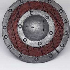May include: A round, wooden shield with a metal rim and a central metal boss. The shield is painted in shades of red and silver, with a wood grain pattern visible on the surface.
