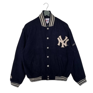 Può includere: Una giacca da baseball in lana blu navy con collo e polsini a righe bianche e nere. La giacca presenta un grande logo ricamato dei New York Yankees sul retro e un logo più piccolo sulla manica sinistra. La giacca ha una chiusura a bottoni e due tasche.