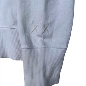 以下が含まれることがあります： 左袖にリブの袖口と小さな刺繍の「XX」ロゴが付いた白い長袖スウェットシャツです。