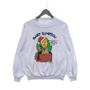 Może przedstawiać: Biała bluza z nadrukiem Bart Simpson w czapce Świętego Mikołaja trzymającego prezent. Tekst "Bart Simpson" jest nadrukowany w niebieskim kółku nad grafiką. Tekst "Wesołych Świąt, człowieku!" jest nadrukowany w dymku obok grafiki.
