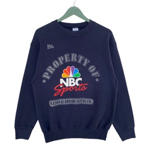 May include: A navy blue sweatshirt with a large graphic print of the NBC Sports logo. The logo features the iconic peacock design with the words "Property of NBC Sports National Broadcasting Co."