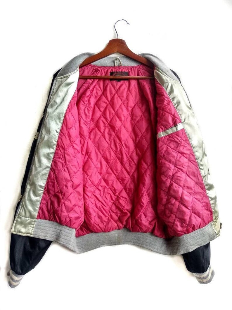 May include: A black and silver satin bomber jacket with a pink quilted lining. The jacket has a grey ribbed collar, cuffs, and waistband.
