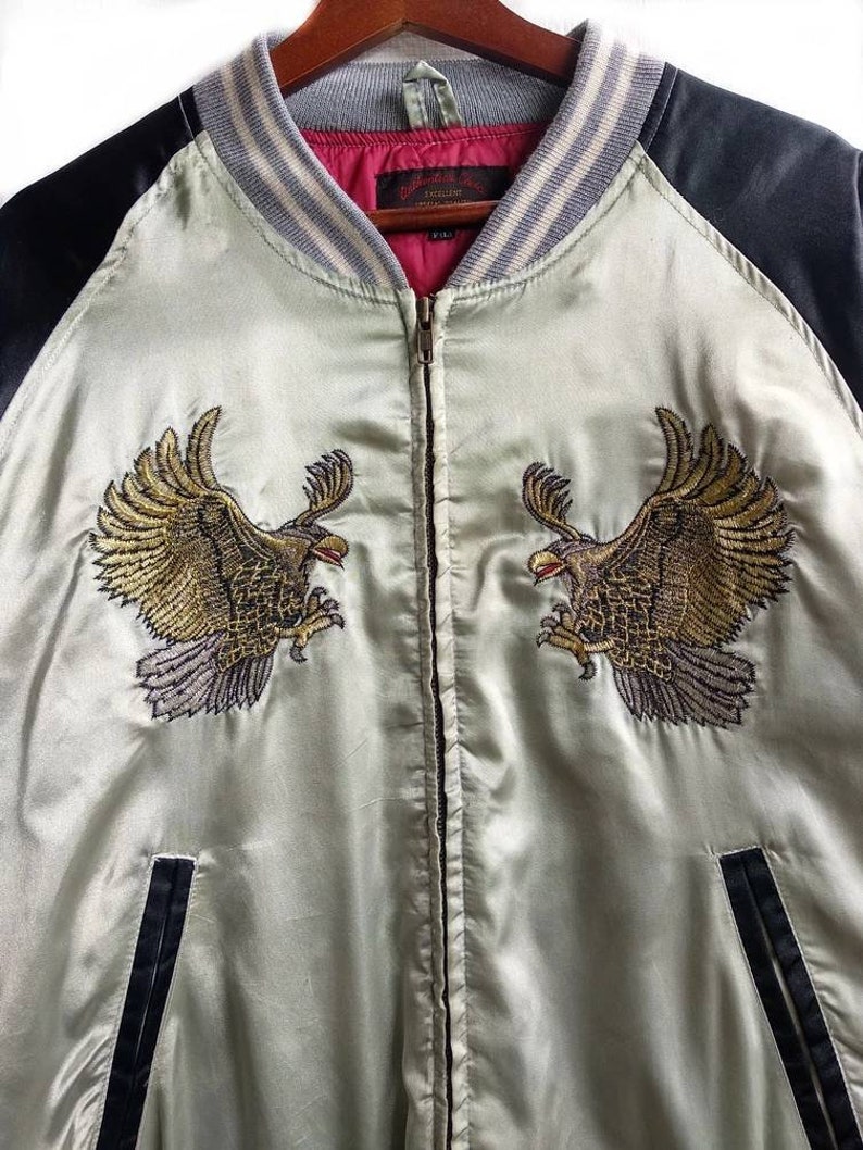 May include: A light green satin bomber jacket with black sleeves and a zipper closure. The jacket features embroidered golden eagles on the front and a red and gray striped collar.