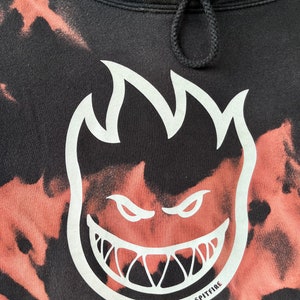 May include: Black tie-dye hoodie with a white Spitfire logo featuring a flame and a grinning face. The logo says "Spitfire" at the bottom.