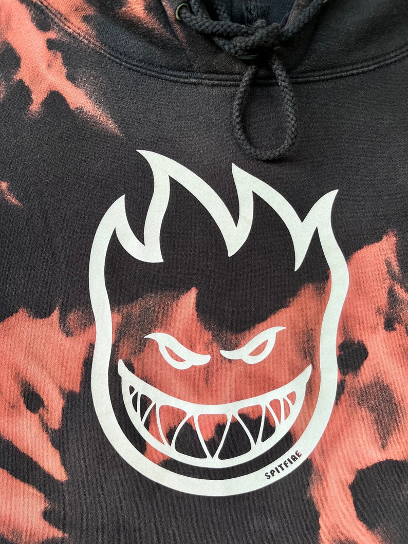 May include: Black tie-dye hoodie with a white Spitfire logo featuring a flame and a grinning face. The logo says "Spitfire" at the bottom.