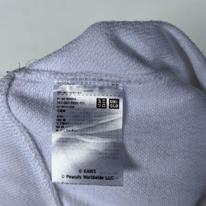 以下が含まれることがあります： 白地に黒文字のタグ。タグには「341-407)38:01-011」「01-26-8H087A」「341-407198191-01)」「62-001A-028」「中国製」「本体」「100%」「リブ部分」「96%」「エポリウレタン」「ユニ UNI」「クロ QLO」「KAWS」「Peanuts Worldwide LLC」の文字が書かれている。