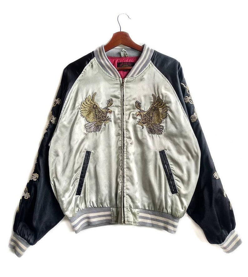 May include: A silver satin bomber jacket with black sleeves and a contrasting gray and white striped trim. The jacket features embroidered golden eagles on the front and intricate floral designs on the sleeves.