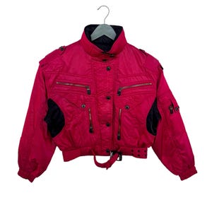 May include: A vintage magenta ski jacket with black accents. The jacket features multiple zippered pockets, a belted waist, and a high collar. The jacket is hanging on a silver hanger.