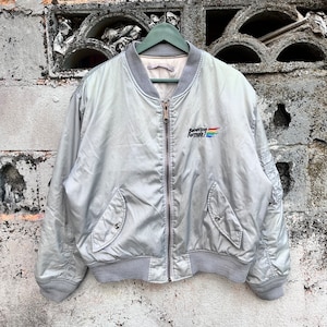 May include: A silver bomber jacket with a Benetton Formula 1 logo on the left chest. The jacket has a zipper closure and two pockets.