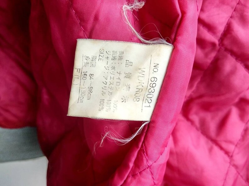 May include: A white tag with black text detailing the size, fabric composition, and care instructions for a jacket. The tag reads "NO. 693021 WDKROB Quality Display Outer Fabric: Nylon 100% Lining: Polyester 100% Jersey: Acrylic 100% SIZE Chest 84-98cm Height: 160-180cm F(L)"
