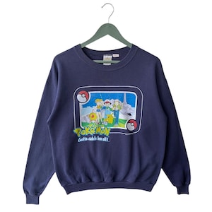 May include: Navy blue sweatshirt featuring a colorful graphic of Pokémon characters within a framed design. The graphic includes the text "Pokémon" and "Gotta catch 'em all!" The sweatshirt is hanging on a green hanger.