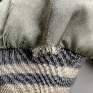May include: Close-up of a green satin bomber jacket with a gray and white striped knit cuff. The satin fabric has a slight sheen and the knit cuff is soft and textured.