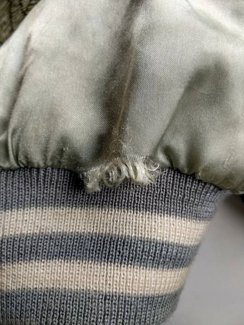 May include: Close-up of a green satin bomber jacket with a gray and white striped knit cuff. The satin fabric has a slight sheen and the knit cuff is soft and textured.